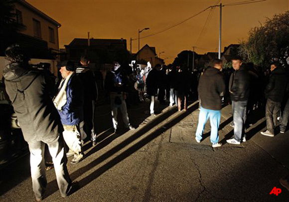 Journalists and residents stand at night next to an apartment building where a suspect in the shooting at the Ozar Hatorah Jewish school is still barricaded in Toulouse, southern France, early Thursday, March 22, 2012. Riot police set off explosions outside the apartment building early Thursday in an effort to force the surrender of the gunman. (AP Photo/Christophe Ena)