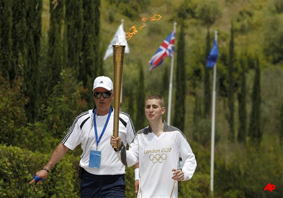 Second Olympic Torch bearer, Alex Loukos from Great Britain carries the flame inside the tomb of Pierre de Coubertin, in front of the EU, British and the Olympic flag following an Olympic flame ceremony on Thursday, May 10, 2012, in Ancient Olympia, Greece. The flame lit in the birthplace of the Ancient Olympics will travel to London, where the Summer Games will take place from July 27 Aug. 12. (AP Photo/Petros Giannakouris)